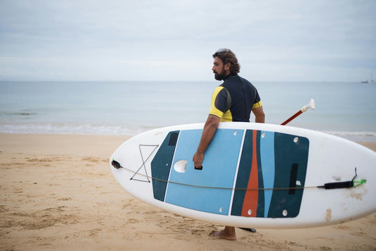 Man carrying a paddleboard on the beach, showcasing summer fitness tips 2025 for active lifestyles